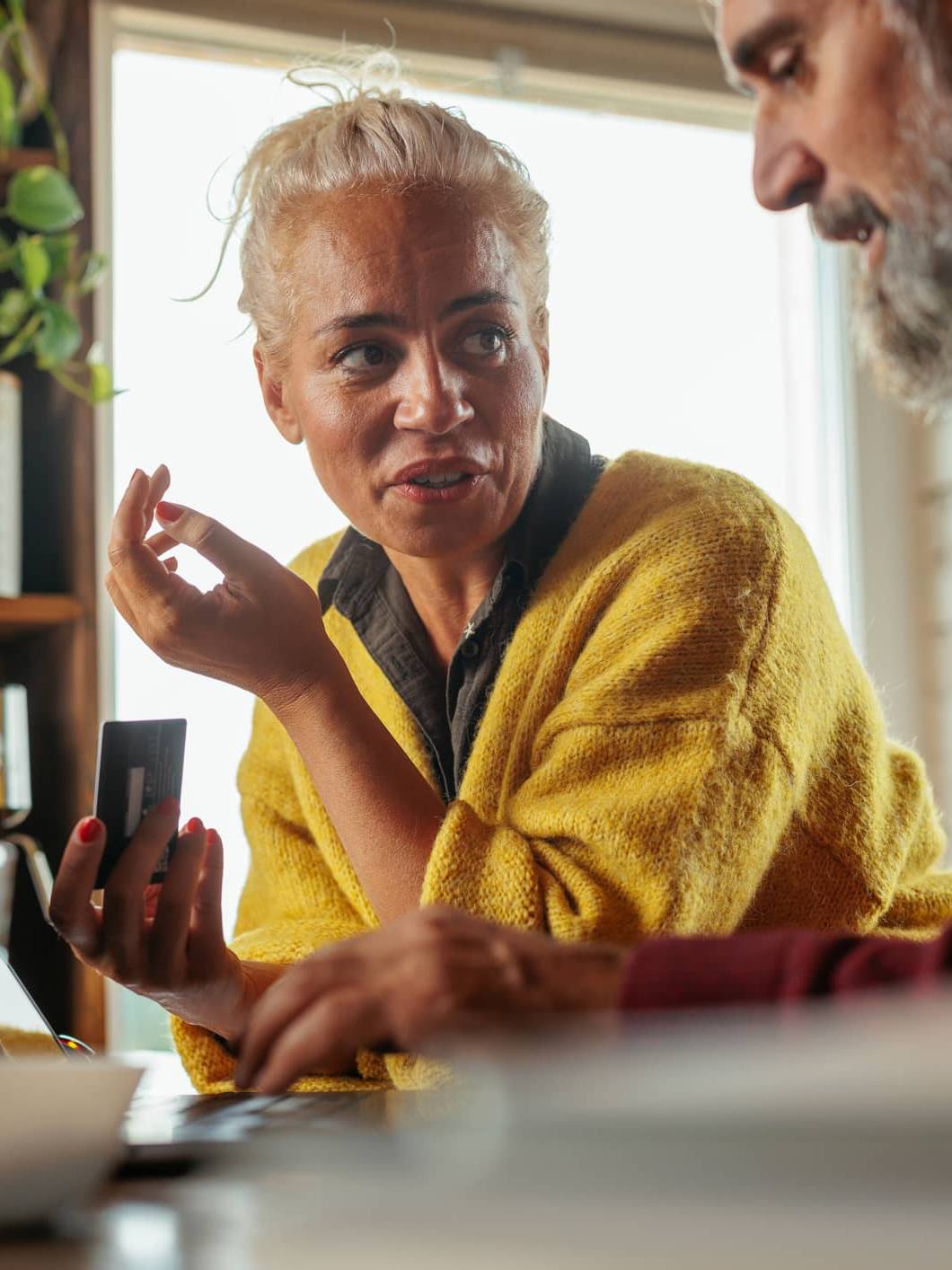 Woman holding a bank card while discussing investments with her partner beside a laptop at home.