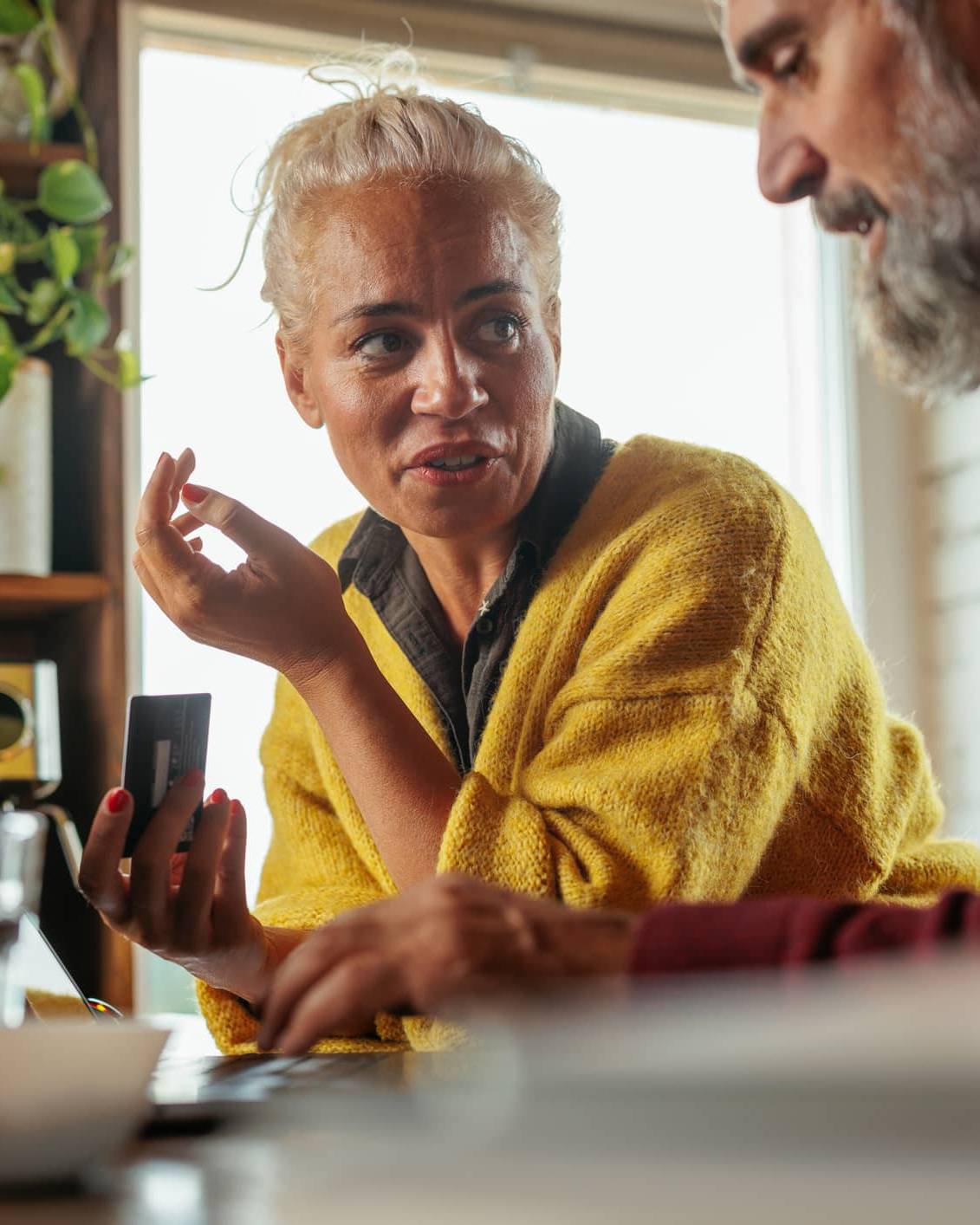 Woman holding a bank card while discussing investments with her partner beside a laptop at home.
