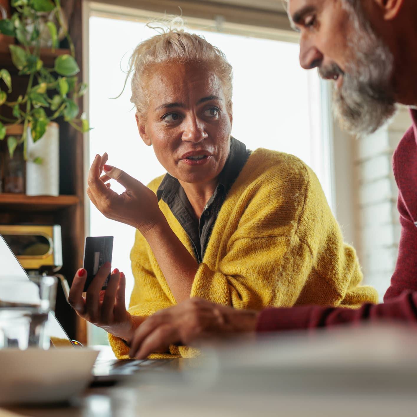 Woman holding a bank card while discussing investments with her partner beside a laptop at home.
