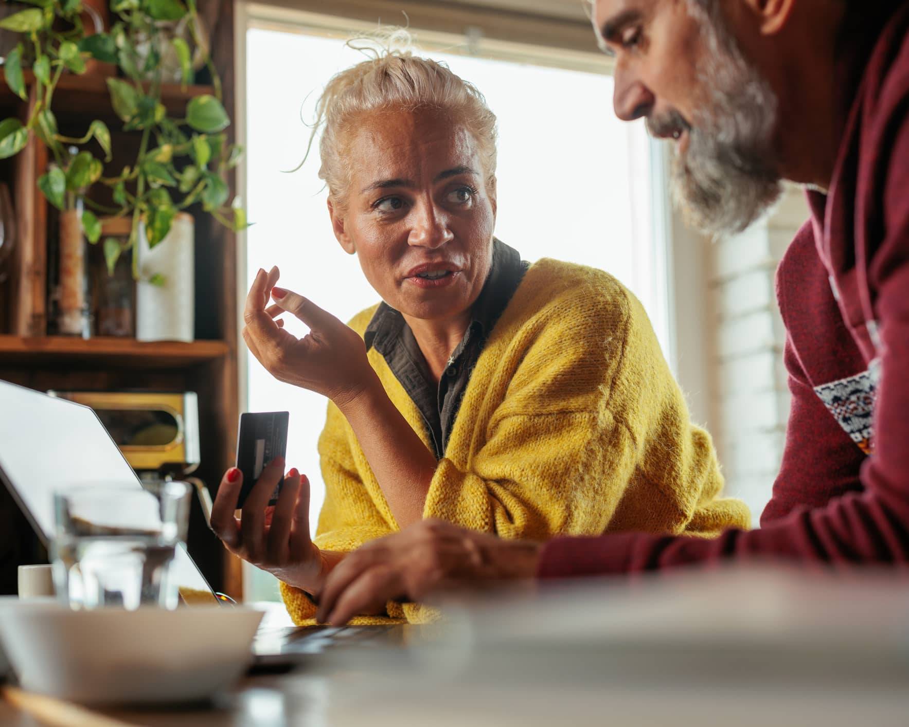Woman holding a bank card while discussing investments with her partner beside a laptop at home.