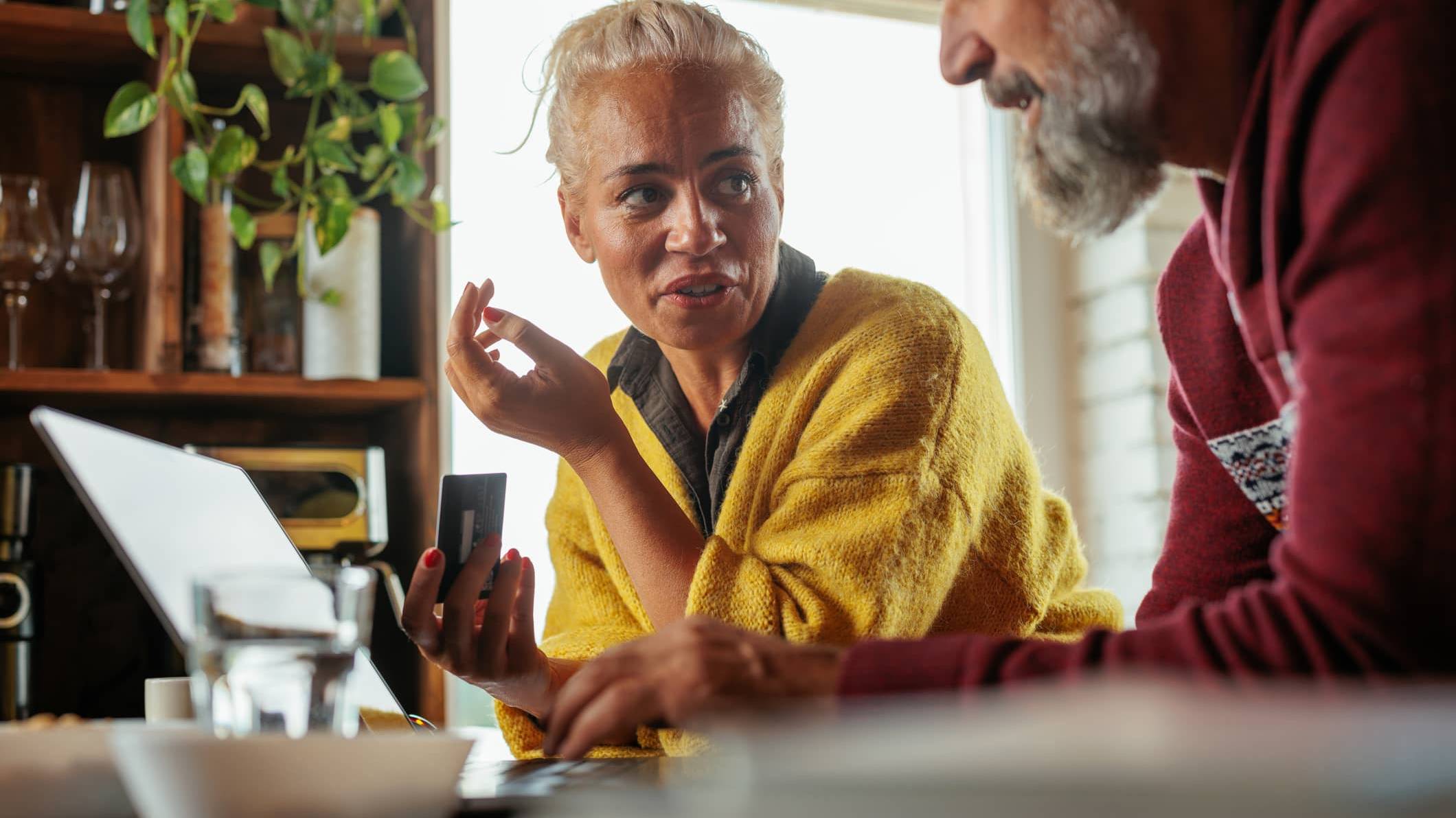 Woman holding a bank card while discussing investments with her partner beside a laptop at home.