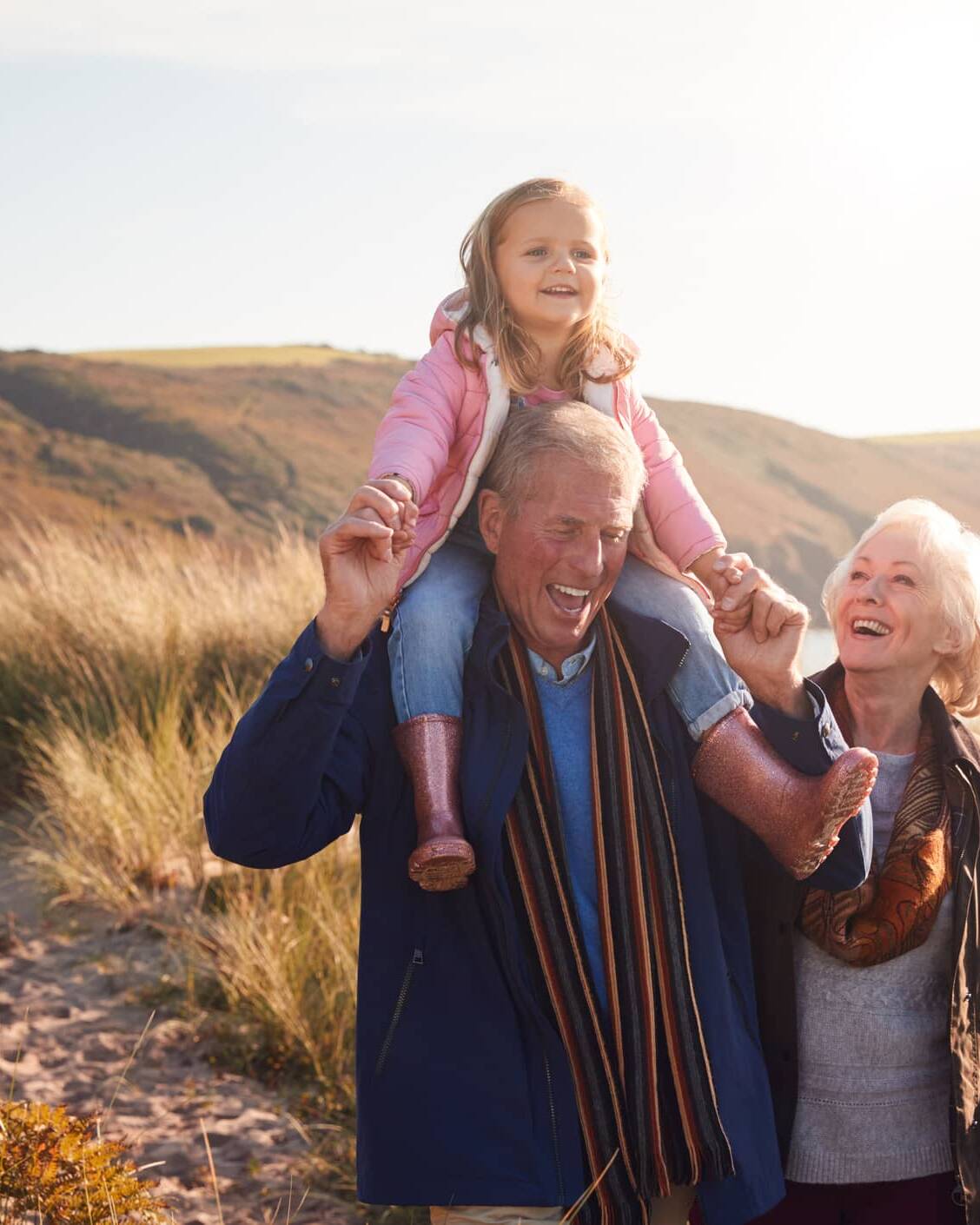 Grandparents enjoying a coastal walk with their young granddaughter sitting on her grandfather's shoulders.