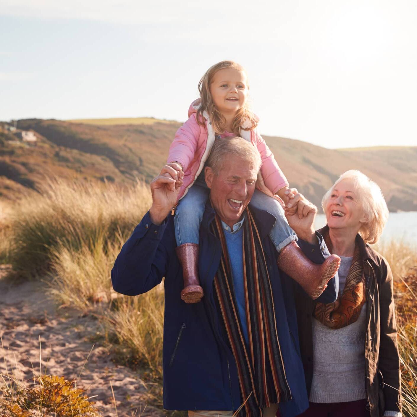 Grandparents enjoying a coastal walk with their young granddaughter sitting on her grandfather's shoulders.