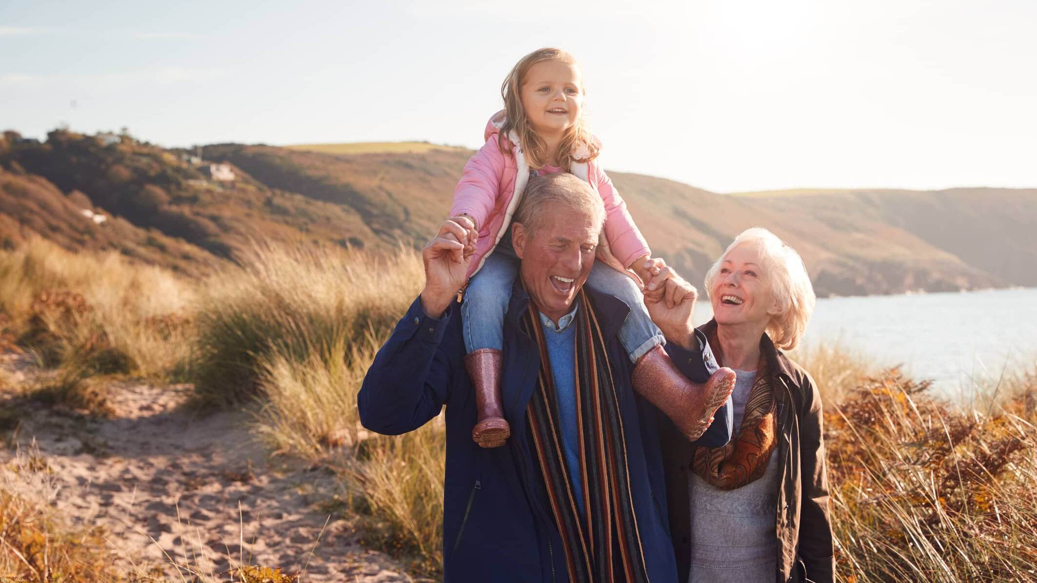 Grandparents enjoying a coastal walk with their young granddaughter sitting on her grandfather's shoulders.
