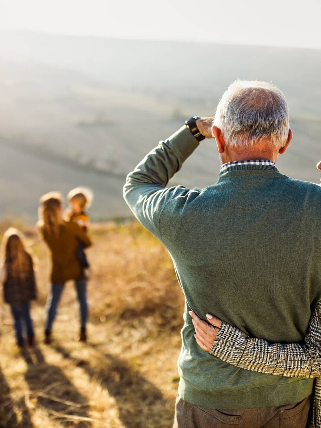 Older couple embracing while watching younger generations of their family walking in the countryside