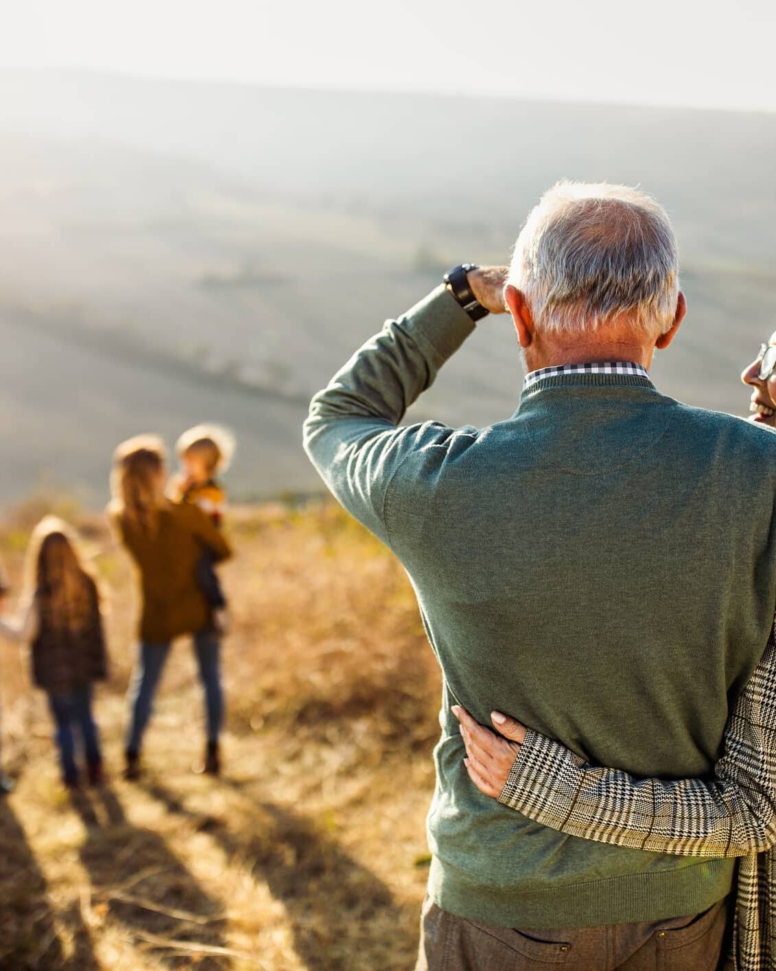 Older couple embracing while watching younger generations of their family walking in the countryside