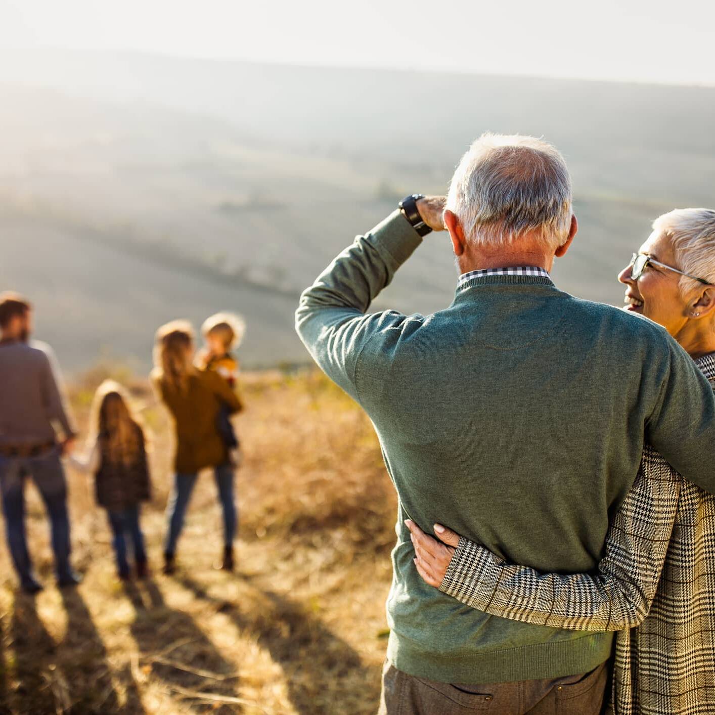 Older couple embracing while watching younger generations of their family walking in the countryside