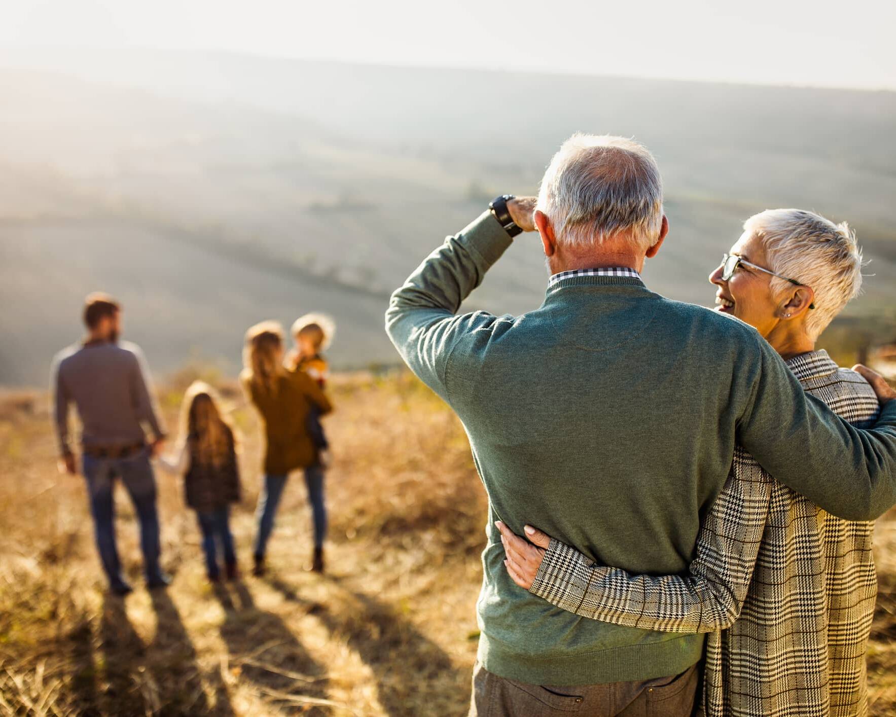 Older couple embracing while watching younger generations of their family walking in the countryside