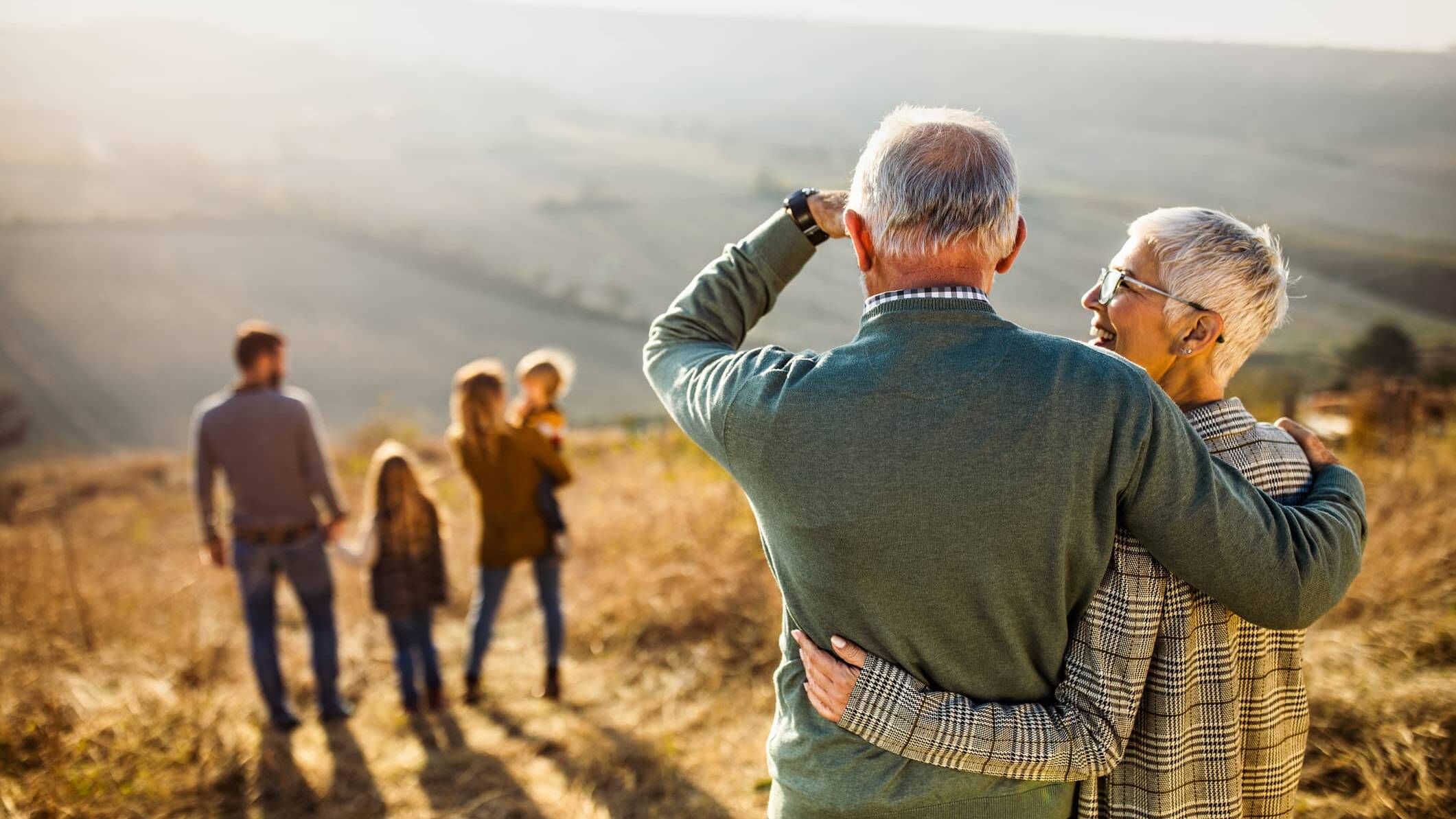 Older couple embracing while watching younger generations of their family walking in the countryside