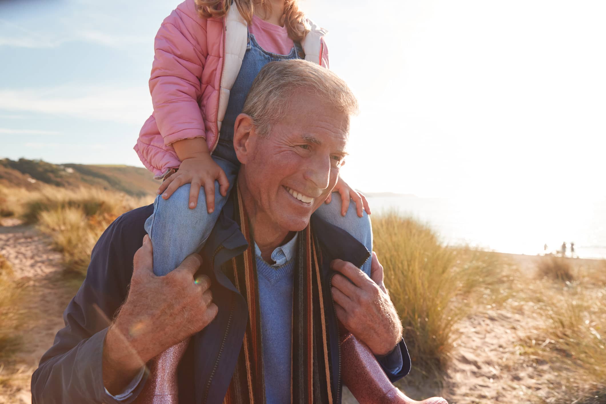 Smiling grandfather carrying his young granddaughter on his shoulders during a walk by the coast.