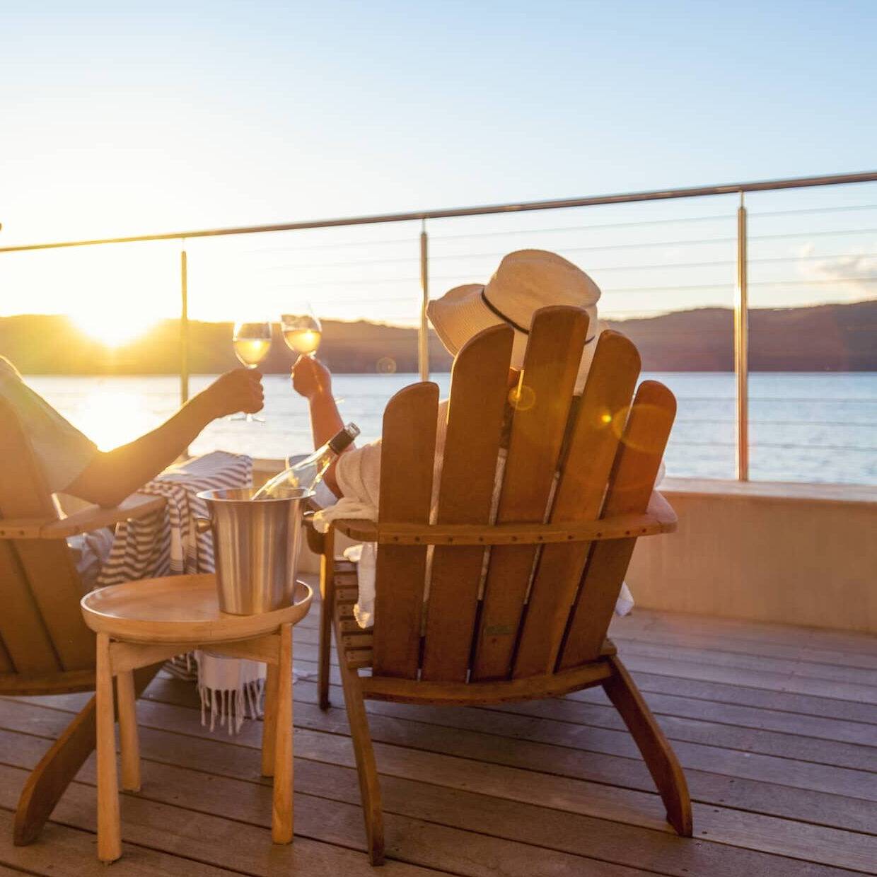 Retired couple relaxing in wooden chairs, raising glasses of wine while watching the sunset over the sea.
