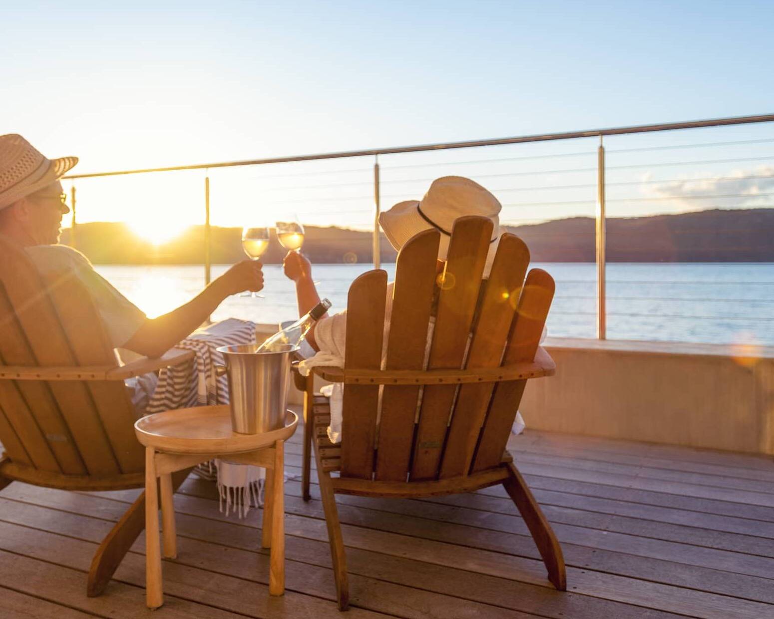 Retired couple relaxing in wooden chairs, raising glasses of wine while watching the sunset over the sea.