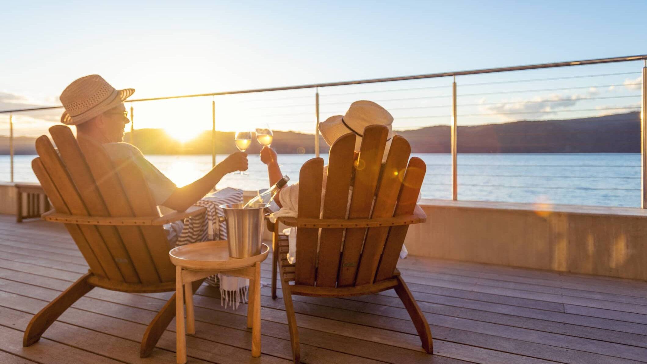 Retired couple relaxing in wooden chairs, raising glasses of wine while watching the sunset over the sea.