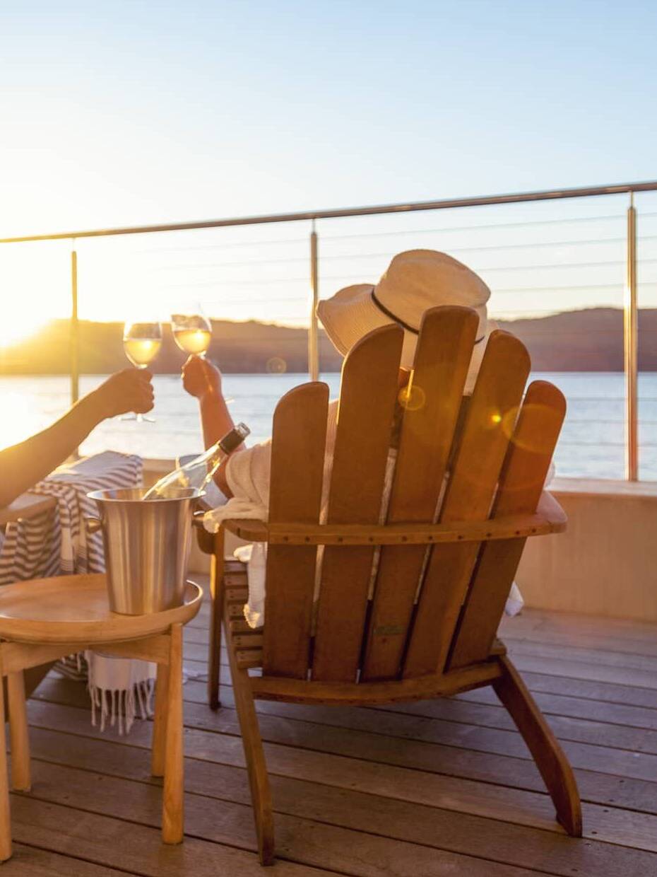 Retired couple relaxing in wooden chairs, raising glasses of wine while watching the sunset over the sea.