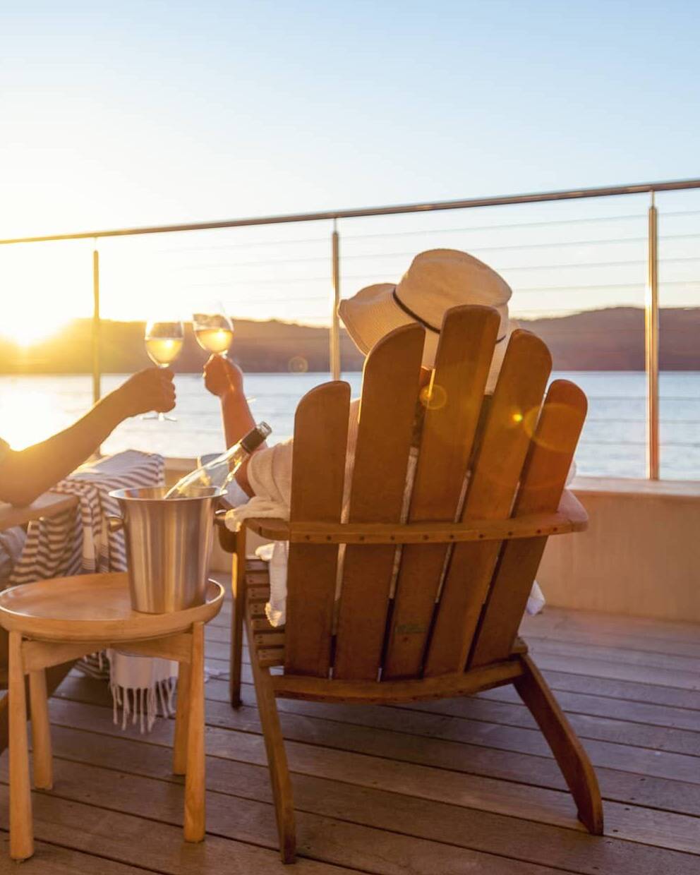 Retired couple relaxing in wooden chairs, raising glasses of wine while watching the sunset over the sea.