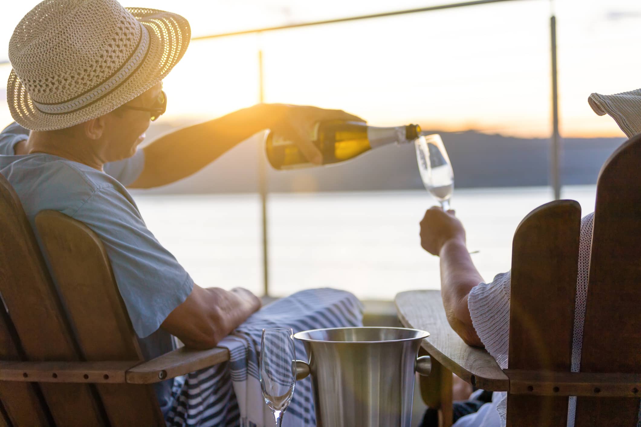 Older couple seated beside the sea at sunset, with one pouring sparkling wine into the other's glass.